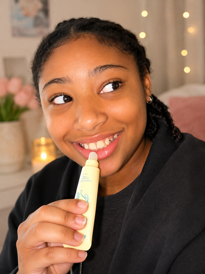 A smiling girl holding a Truly Yours Vanilla Butter lip balm tube with blue lettering, set against a softly blurred indoor background with warm lights and pink flowers.
